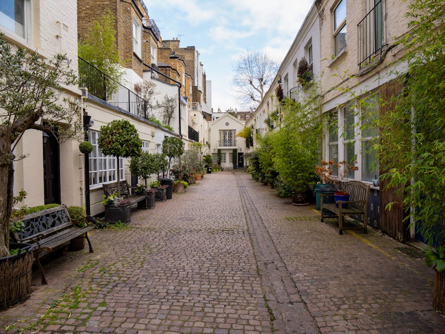 A narrow cobblestone alleyway in a residential area, flanked by white-painted townhouses with large windows and small balconies decorated with potted plants. Several benches and a variety of potted greenery, including small trees and shrubs, are positioned along the front of the buildings on both sides. The alley is lined with narrow, uneven cobblestones and shows signs of minimal wear, with some patches of moss or grass between the stones. In the background, there is a small, white, two-storey building with a central doorway and large windows, set against a partly cloudy sky. The environment appears calm and well-maintained, with a mix of natural greenery and human-made structures, typical of an urban residential courtyard or communal outdoor space, which may be a setting where private rubbish clearance services, like those offered by Rubbish Collection Kensington, are often employed for maintaining cleanliness and tidiness.