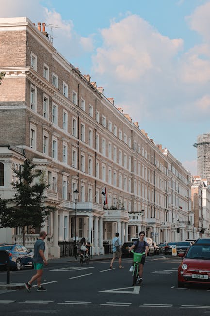 A street scene in Kensington featuring a row of multi-story residential buildings with classic architectural details, including large windows, decorative cornices, and stone façade finishes in light beige and white tones. The buildings are closely spaced, with some small trees and black lampposts positioned along the pavement in front. In the foreground, several people are visible, including a cyclist in a black shirt and green shorts, a pedestrian in a gray top and beige pants, and others walking along the sidewalk. Parked cars, including a red vehicle on the right and a dark-colored one further back, line the street. The sky is partly cloudy with patches of blue, and a construction crane is visible in the distance, indicating ongoing development nearby. The scene exemplifies an urban environment where private rubbish collection or on-site clearance may be necessary, supported by the context of local waste management services like those provided by Rubbish Collection Kensington.