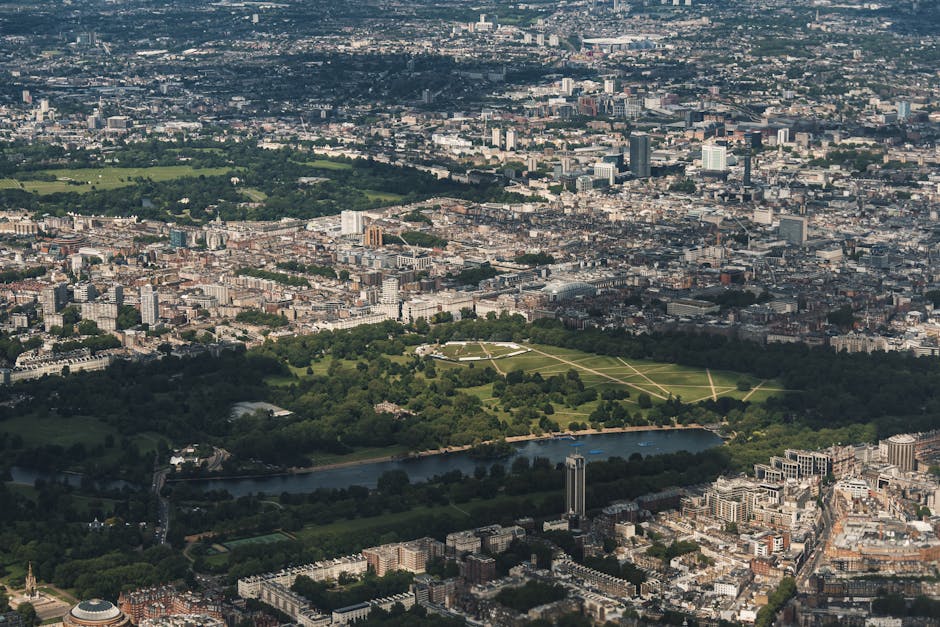 An aerial view of central London featuring a large rectangular park with expansive green lawns, pathways crossing diagonally and in grid patterns, surrounded by dense urban development with numerous multi-storey buildings of varying heights. Trees border the park, and a calm river flows along its southern edge, lined with additional green spaces and pathways. The skyline includes mid-rise and high-rise buildings, with some taller structures in the distance, under a slightly cloudy sky. The overall landscape showcases a mix of natural and constructed elements, reflecting the urban environment's integration of open parks within the cityscape, suitable for visualizing the context of alternative waste handling or private disposal options nearby.