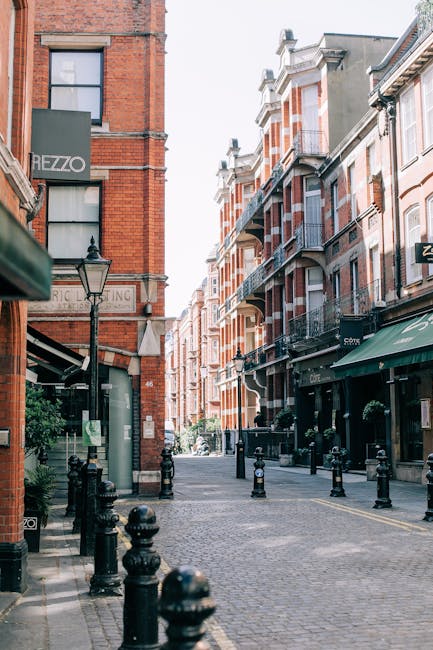 A narrow cobbled street lined with red-brick Victorian-style buildings featuring white window frames and decorative iron balconies. On the left, there is a building with a black sign reading 'HEZZO,' and a traditional lamppost with a glass lantern is positioned beside it. The street is bordered by black metal bollards separating the pedestrian area from the roadway, which is free of vehicles. Several hanging plant pots with greenery are suspended from the building facades on the right side, alongside a storefront with a green awning. The overall scene is well-lit with natural daylight, creating a clean and tidy urban atmosphere. This setting reflects an alternative approach to waste management, typical of private rubbish collection services operating within a cityscape, with no visible rubbish or waste debris in the vicinity, emphasizing the area’s neatness and cleanliness that such services aim to maintain as part of maintaining an organized environment.