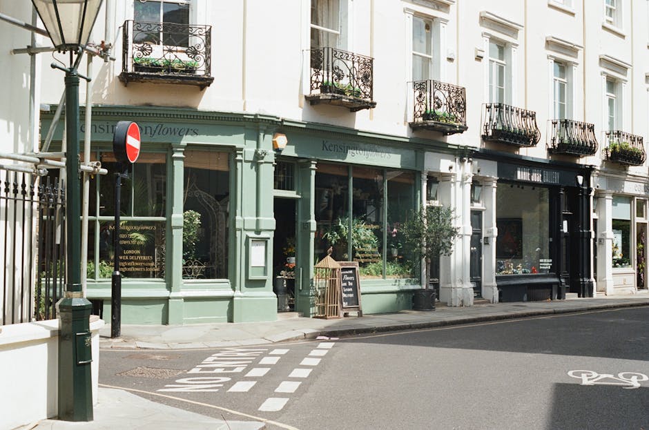 The image depicts a quiet street scene in front of a flower shop located on the ground floor of a white, multi-storey building with large windows. The shop’s facade is painted in a soft green color with decorative moldings, and its large glass windows reveal an assortment of potted plants, floral arrangements, and garden supplies inside. A small wooden A-frame chalkboard sign is positioned on the sidewalk outside the shop, advertising its offerings. To the left, a vintage-style street lamp stands near a black metal gate that encloses a small area, possibly a side entrance, with a no-entry traffic sign mounted beside it. The street features a dedicated cycle lane marked with white line and bike symbol, and a pedestrian crosswalk with bold white stripes runs across the asphalt road in front of the shop. Above, several narrow balconies with black wrought iron railings and potted plants extend from the upper floors, adding a charming architectural detail. The scene appears to be well-lit with natural daylight, creating a calm and inviting atmosphere, relevant to local waste management and rubbish removal services that may support maintaining such urban environments.