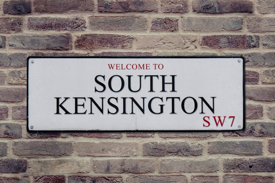 A rectangular metal street sign mounted on a brick wall, displaying the text 'Welcome to South Kensington' with the postcode 'SW7' in the lower right corner. The sign has a white background with black lettering, and the words 'Welcome to' and 'SW7' are in red. The brick wall behind the sign consists of reddish-brown and beige bricks arranged in a traditional horizontal pattern, with visible mortar lines. The sign is secured with four small screws, one in each corner. The environment appears to be an exterior urban setting, typical of a residential or commercial area in South Kensington, London. The lighting is natural, providing clear visibility of both the sign and the textured brick surface, making it suitable for use in accessibility-focused content about London locations or local signage.