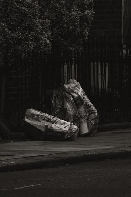 Two large black rubbish bags made of thick plastic material are placed on a paved sidewalk near a black metal fence, which encloses a garden area with dense foliage and trees. The bags appear to be filled with waste, possibly household or garden rubbish, and are situated close to the curb on the street. The environment is outdoors, with ambient lighting suggesting the scene might have been captured during the late afternoon or early evening. The bags are leaning against each other and the fence, with no visible signage or labels. The setting indicates a private property or a designated spot for rubbish collection, typical for municipal or private waste disposal services. This scene reflects an example of on-site clearance or alternative waste handling, potentially managed by a waste management company such as Rubbish Collection Kensington, known for providing rubbish removal services in residential areas like Kensington.