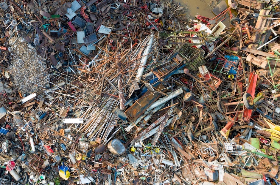 An extensive pile of mixed discarded items and scrap metal, predominantly consisting of rusted iron and steel components, stacked outdoors on uneven ground. The debris includes broken bicycle frames, twisted metal rods, rusted pipes, and various metal parts with textured, weathered finishes. Surrounding the tangled mass are scattered smaller objects such as plastic and rubber components, with some items partially buried beneath others. In the background, there are hints of muddy water and some vegetation, indicating the waste is situated in a neglected or industrial outdoor area. The overall scene is illuminated naturally, highlighting the corrosion, discoloration, and complex textures of the collected scrap. This image exemplifies the type of waste material often managed by private rubbish removal services like Rubbish Collection Kensington, which handle large, bulky, and metal-heavy waste streams that may be incompatible with standard municipal disposal methods, illustrating the necessity for specialized collection and recycling solutions for metal and mixed waste.
