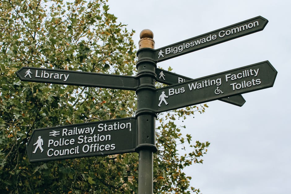 A black multi-directional pedestrian signpost outdoors, mounted on a cylindrical pole with a rounded top, featuring six rectangular signs with white text and walking icons. The signs point in different directions towards locations such as 'Biggleswade Common,' 'Library,' 'Bus Waiting Facility Toilets,' 'Railway Station,' 'Police Station,' and 'Council Offices,' with the Railway Station sign including a rail icon. The background shows a large leafy tree with green and yellowish leaves, and the sky is overcast with pale grey clouds, creating even, diffuse lighting. The signpost is situated on a paved area, possibly a town center or park, and the overall scene exemplifies outdoor signage for navigation and local facilities, which is relevant to alternative waste handling or public service environments that may be managed by private providers like Rubbish Collection Kensington for private waste removal services.