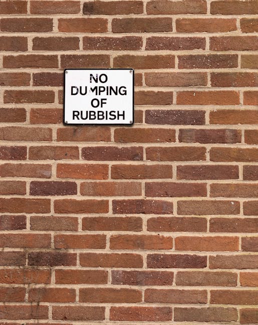 A white rectangular metal sign mounted on a red brick wall, displaying the message 'NO DUMPING OF RUBBISH' in bold black uppercase letters. The sign is positioned slightly to the upper left of the image, attached with four screws, one at each corner. The brick wall behind the sign consists of uniformly laid bricks with a reddish-brown hue, some darker and some lighter, with light gray mortar joints creating a regular horizontal pattern. The bricks have a slightly rough texture, typical of traditional brickwork. The lighting appears natural, casting minimal shadows, and the overall scene is neutral and professional, fitting within an environment that emphasizes community rules regarding waste disposal. The presence of this sign suggests a location where private or independent waste management, such as from rubbish removal services like Rubbish Collection Kensington, is applicable, reinforcing the context of controlled rubbish handling and the importance of adhering to local regulations.