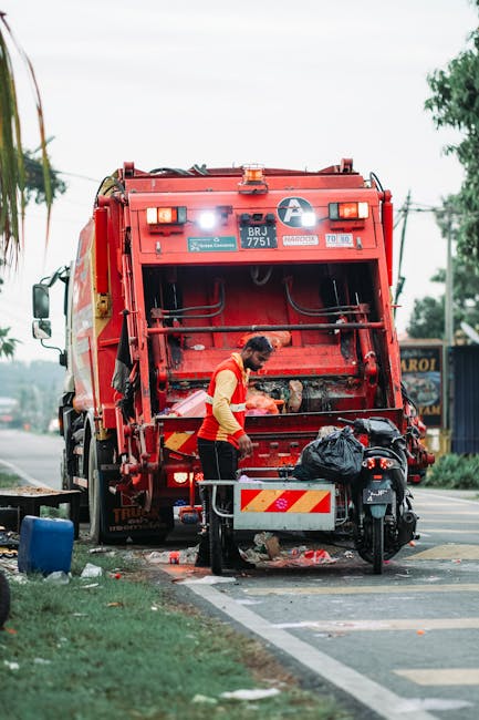 A waste collection worker wearing a red and yellow high-visibility uniform is emptying a black trash bag into the open rear hopper of a large red rubbish collection truck parked on the side of a street. The truck features safety and identification labels, with the license plate number 'BRJ 7751' visible on the back. The scene is set during daytime, with a cloudy sky above and a background consisting of trees, utility poles, and a blue street-side building or shop. The area around the truck includes a small blue container, a motorcycle parked nearby with a side bag, and some scattered debris or rubbish on the pavement, indicating a typical urban waste collection context. The setting suggests an on-site or private rubbish removal operation, consistent with alternative waste handling services provided by companies like Rubbish Collection Kensington.