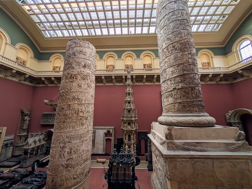Inside an ornate historical building with high ceilings and large skylights, two massive ancient marble columns with detailed, weathered surfaces flank the scene, standing on elevated rectangular bases. Behind the columns, a decorative, vertically-tiered marble sculpture or column fragment extends upward, showcasing intricate carvings and a layered, sculpted design. To the left, smaller display cases and artifacts are visible, suggesting a museum or gallery environment. The walls are painted deep pink, contrasting with the light-colored marble of the historic objects. The upper balcony features classical balustrades and decorative arches, illuminated by natural light entering through arched windows and the skylight ceiling above. The scene emphasizes the grandeur of the classical antiquities, with a neutral and professional tone fitting an exhibition of historical artifacts, subtly aligned with a focus on private collection or preservation of ancient ruins and architectural remains.