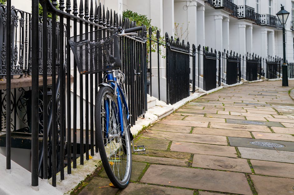 In the foreground, a blue bicycle with a black plastic front basket is leaning against a black wrought iron fence that lines a sidewalk in a residential area. The bicycle's frame is metallic blue with silver spokes on the front wheel, and it has black tires and pedals. The fence features pointed finials and is positioned alongside white stone steps leading up to a row of terraced terraced houses. Behind the fence, partial views of the building’s white facade and black wrought iron balconies are visible. The sidewalk consists of large, irregularly shaped paving stones in shades of brown and beige, with narrow gaps of moss growing between them. The scene is evenly lit, suggesting natural daylight without strong shadows. The overall environment appears clean and well-maintained, with the fencing and building details providing an elegant urban residential context, subtly relevant to waste and rubbish collection practices on private property or property boundaries, as seen in professional rubbish removal services. The image is captured in a manner aligning with visually accurate descriptions for accessibility and SEO relevance related to rubbish collection services in urban settings.