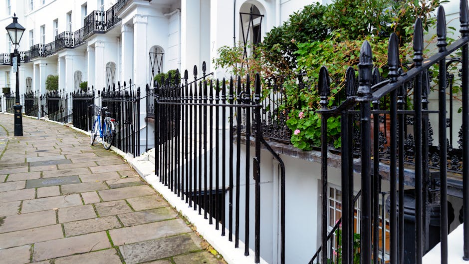 A row of white terraced houses with black wrought iron railings along a paved sidewalk in Kensington. In the foreground, a blue bicycle leans against the railing near a small white basement window. The railings feature pointed finials and are positioned in front of a garden area with green foliage and pink flowering plants. The sidewalk consists of large, weathered stone slabs with uneven edges, leading toward the background, where more similar houses are visible. The exterior walls of the houses are smooth and painted white, with black metal window frames and decorative street lamps mounted on the façades. The lighting appears natural and diffused, suggesting an overcast day. This scene exemplifies typical residential street architecture, often requiring private rubbish collection services like those provided by Rubbish Collection Kensington, especially for on-site waste clearance or alternative waste handling in the area.