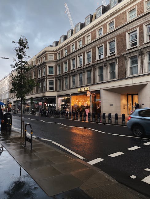 A street scene in an urban area showcasing a multi-story building with a light brown brick facade and numerous evenly spaced sash windows. The ground floor features a commercial unit with a brightly lit interior, displaying orange and yellow signage and seating visible through large glass windows. In front of the building, the pavement is wet and reflective, with a row of black bollards separating the pedestrian walkway from the roadway. A silver car is seen passing by, and a few pedestrians are near the entrance of the commercial establishment. To the left, a small tree with dark green leaves partially obscures the view of additional storefronts further along the street. The sky above is overcast, contributing to the subdued lighting. The scene depicts typical city commercial and residential surroundings, with an emphasis on urban waste management and alternative rubbish collection options, such as private waste handling for businesses and buildings in the area, consistent with services offered by Rubbish Collection Kensington.