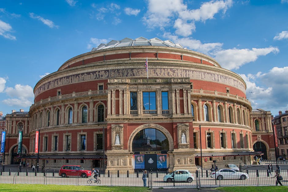 A large, historic round building with a domed glass roof, constructed from red brick and stone details. The facade features multiple arched windows on the lower level, framed by ornate stonework, with rectangular windows on the upper level, some of which are adorned with small statues in niches. An entrance archway is centered at the front, displaying a sign that reads 'Royal Albert Hall,' with a Union Jack flag flying above it. The building is situated on a paved area with a black metal fence, and in the foreground, there are several vehicles, including a red van and a white car, parked along the street. Pedestrians and a cyclist are visible on the sidewalk. The sky above is bright blue with scattered white clouds, and surrounding urban buildings are visible in the background, highlighting the building’s location in a busy city environment, consistent with a setting where private rubbish collection services might be employed for waste removal after events or maintenance activities at the Royal Albert Hall.
