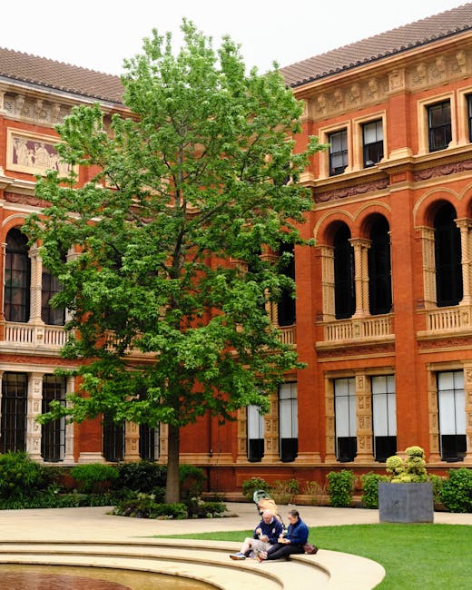 In front of a historic red-brick building with ornate architectural details and large arched windows, a tall deciduous tree with bright green foliage occupies most of the scene. The tree's branches extend outward, partially obscuring the upper floors, while its trunk stands firmly on the paved area. The building's exterior features decorative stonework, cornices, and black window frames, contrasting with the vibrant greenery. In the foreground, two individuals sit on a curved stone bench surrounded by a well-maintained grassy area and small ornamental shrubs. One person is wearing a navy jacket with light-colored pants, and the other is dressed in a dark uniform, possibly a security or staff member. To the right, a large rectangular planter with a small, rounded shrub adds to the landscape’s organized appearance. The overall scene appears to be in a peaceful courtyard or public space, possibly part of a private estate or cultural institution, with natural elements thoughtfully integrated within the historic setting to facilitate independent waste handling or slight on-site clearance, in line with private rubbish removal services.
